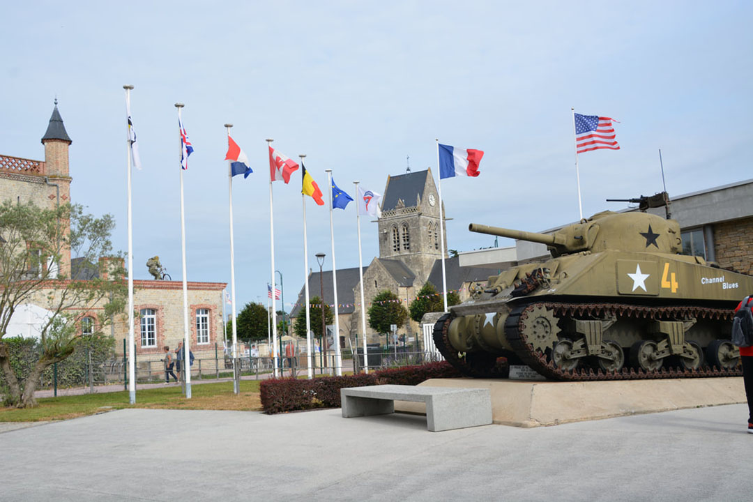 A row of international flags, an old military tank, and Notre Dame de L'Assumption Church at St. Mere Eglise