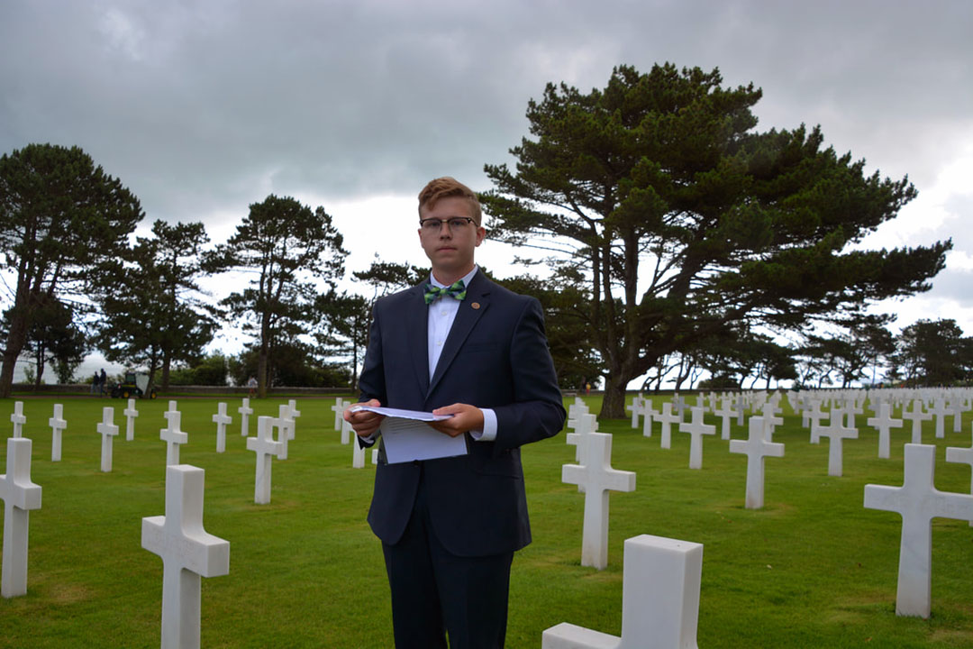 A young person in a suit and tie stands in a field of white crosses holding a paper, ready to give a eulogy