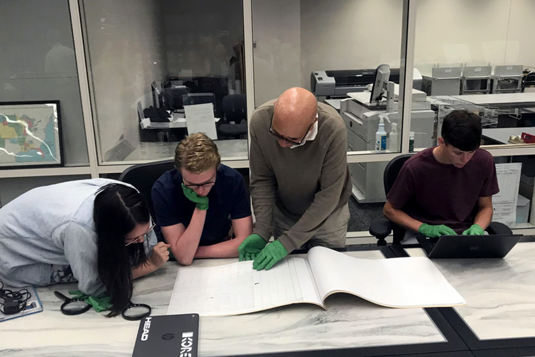 A professor and three students, all wearing gloves, sit together looking at an oversized book in an archive