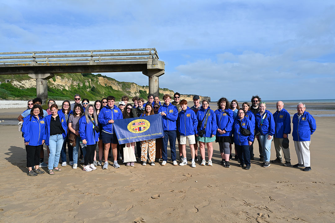 A large group of students in blue jackets standing on Omaha beach on a sunny day