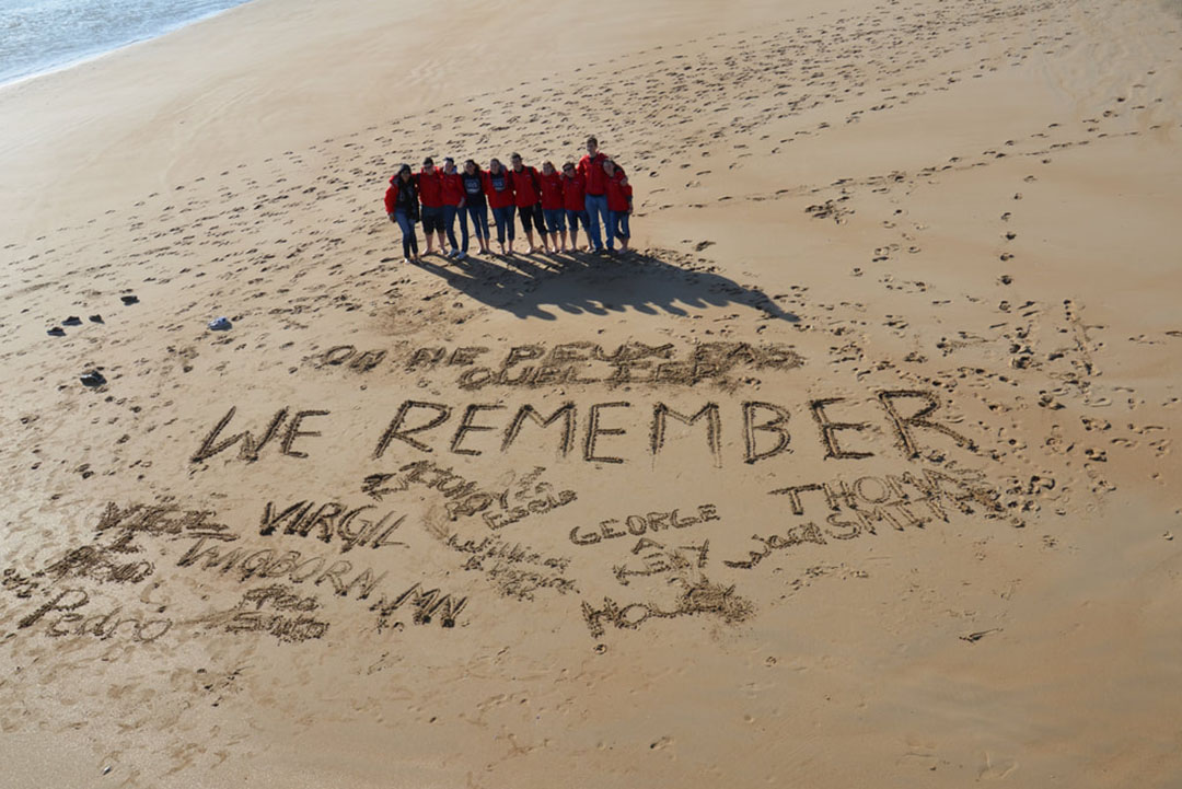 Omaha Beach We Remember A group of people standing on a beach photographed from above, with the words "we remember" traced into the sand, along with people's names