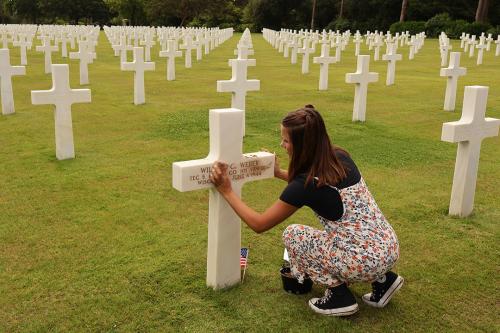A student kneeling in front of a white cross and rubbing sand into the engravement