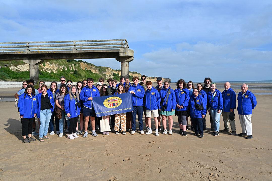 A large group of students in blue jackets standing on Omaha beach on a sunny day